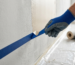 Ein Nahaufnahme-Foto eines professionellen Malers in einem blauen Arbeitshandschuh, der sorgfältig blaues Klebeband von einer strukturierten verputzten Wand entfernt. Eine saubere Linie frischer, halb-trockener beige Farbe ist sichtbar, während das Klebeband einen scharfen Kantenverlauf offenbart.