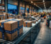 Eine Aufnahme aus einem logistischen Versandzentrum, in dem ein Mitarbeiter im Vordergrund einen Karton mit kältebeständigem Naturkautschuk-Klebeband verschließt, während im Hintergrund Förderbänder und Regale voller Pakete für den Q4-Shipping-Peak vorbereitet werden.